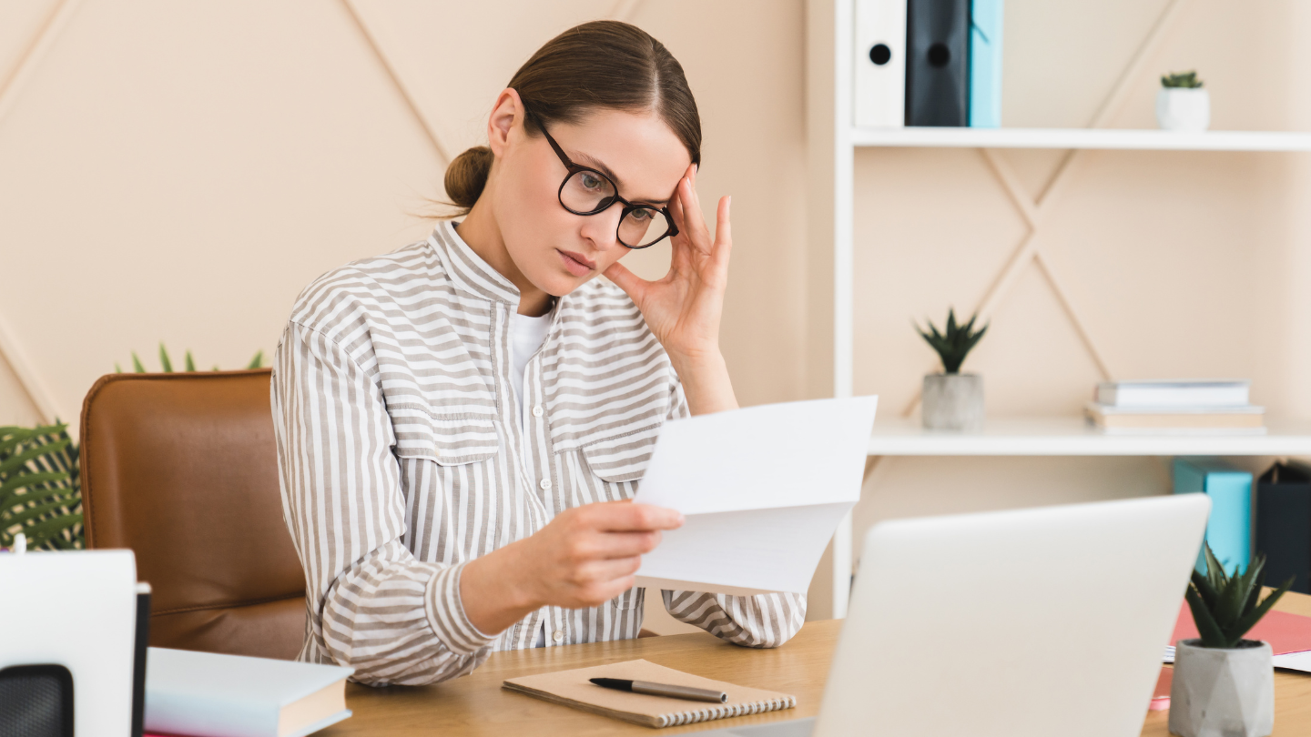 Mujer estresada mirando un documento impreso, sentada al frente de su computador