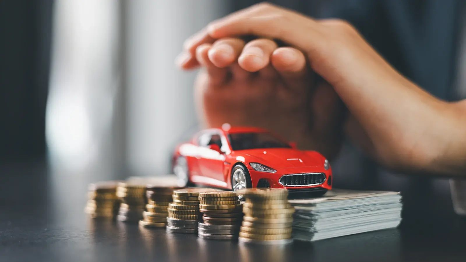 Prenda vehicular. Dos manos, vehículo rojo en miniatura al lado de una fila de monedas